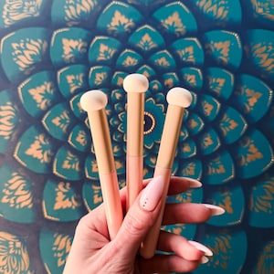 Op de afbeelding: Drie make-upsponsapplicators met beige sponsuiteinden en lichtroze en beige handvatten. De applicators worden vastgehouden voor een turquoise en gouden mandala-patroonachtergrond.