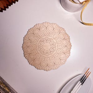 May include: A wooden mandala with a detailed intricate design, perfect for painting or decorating. The mandala is round and has a light brown color.