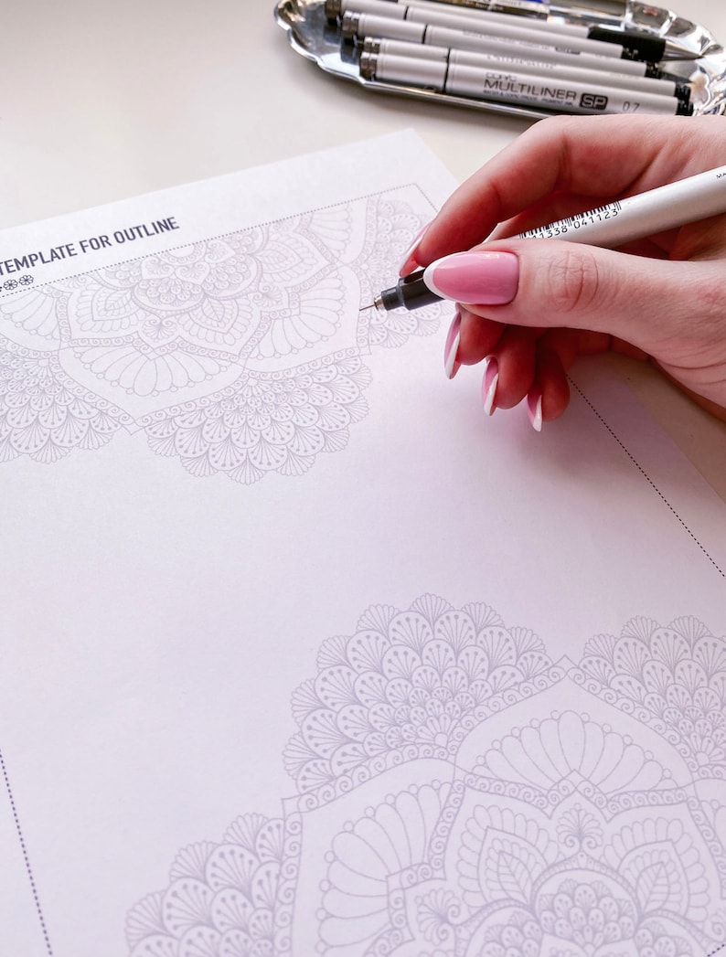 Puede incluir: Una mano sostiene un bol&iacute;grafo blanco y negro y delinea un dise&ntilde;o de mandala detallado en una hoja de papel blanca. El papel tiene el texto "TEMPLATE FOR OUTLINE" en la parte superior.
