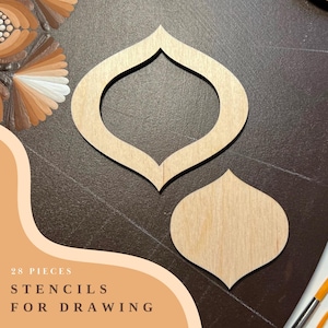 Op de afbeelding: Twee houten stencils in de vorm van traanvormige ornamenten. De stencils liggen op een bruin oppervlak. De tekst "28 PIECES STENCILS FOR DRAWING" is zichtbaar in de linkerbenedenhoek van de afbeelding.
