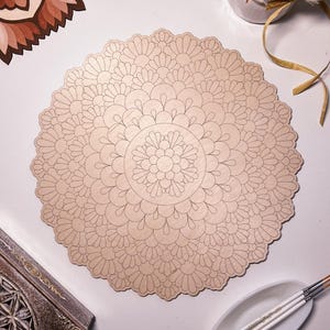Könnte beinhalten: Ein Holzmandala mit einem detaillierten floralen Design, bereit zum Bemalen. Das Mandala ist rund und hat eine hellbraune Farbe. Es ist von Pinseln und einer kleinen Schale mit Wasser umgeben.