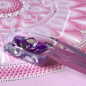 May include: A clear plastic container filled with purple rhinestones and two pink tweezers. The container is sitting on a pink and white mandala pattern.