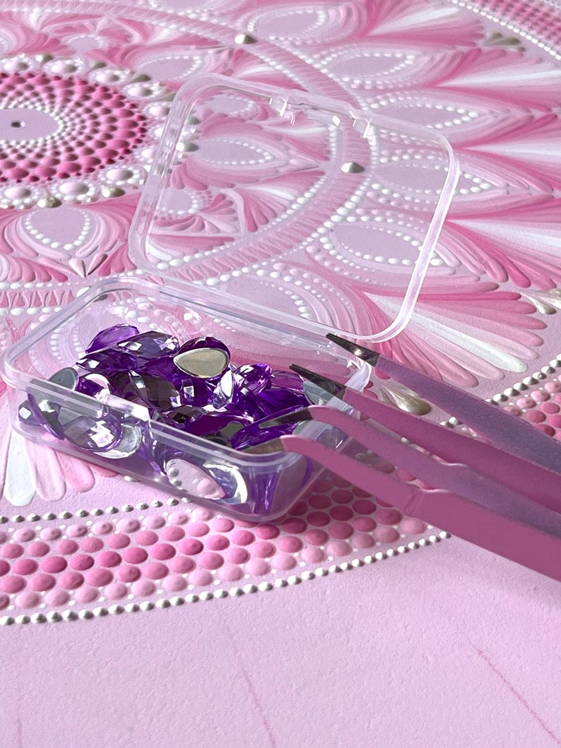 May include: A clear plastic container filled with purple rhinestones and two pink tweezers. The container is sitting on a pink and white mandala pattern.