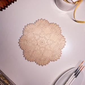 May include: A wooden mandala with a detailed design, ready for painting. The mandala is on a white surface with paintbrushes and a jar of paintbrushes in the background.