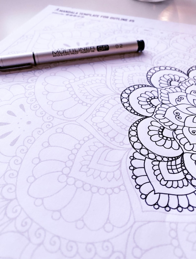 Puede incluir: Un primer plano de un dise&ntilde;o de mandala con detalles intrincados. El dise&ntilde;o est&aacute; parcialmente coloreado con tinta negra. Un bol&iacute;grafo plateado con el texto "MULTILINER SP 02 WATER & COPIC PROOF PIGMENT INK" est&aacute; sobre la p&aacute;gina.
