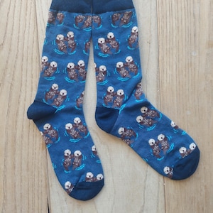 May include: A pair of blue socks featuring a repeating pattern of sea otters in water. The otters are brown and white, and the water is light blue. The socks have dark blue cuffs and toes.