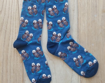 May include: A pair of blue socks with a repeating pattern of sea otters floating in water. The otters are brown and white, and the water is a lighter blue. The socks have dark blue cuffs and toes.