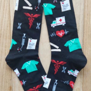 Könnte beinhalten: Schwarze Socken mit medizinischen Symbolen. Das Muster umfasst einen Caduceus, OP-Kleidung, einen Herzmonitor, einen Krankenwagen und Spritzen. Diese Socken sind für Erwachsene konzipiert.
