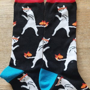 May include: Black socks featuring a repeating pattern of white cats with headbands and fish. The socks have red cuffs and blue heels.