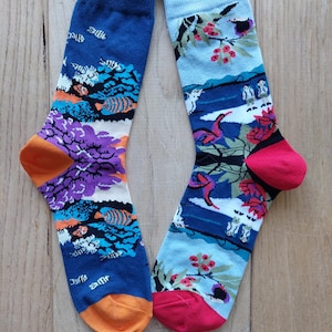 Puede incluir: Un par de calcetines coloridos con diseños únicos. Un calcetín presenta un fondo azul con peces y coral, mientras que el otro tiene un fondo azul claro con pájaros y flores. Ambos calcetines tienen detalles en naranja y rojo.