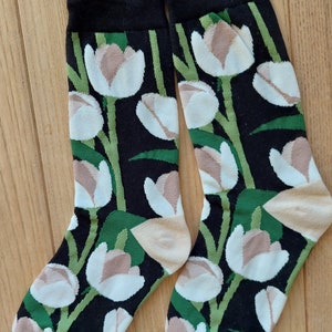 Può includere: Un paio di calzini con sfondo nero e un motivo floreale di tulipani bianchi e beige con steli e foglie verdi. Il bordo superiore è nero, il tallone e la punta sono beige e verdi.