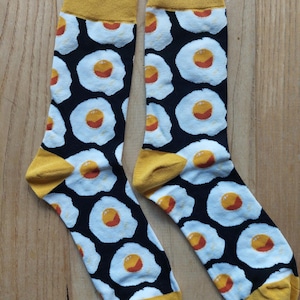 May include: A pair of black socks with a white and yellow fried egg pattern. The socks have a yellow cuff.