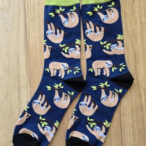 May include: A pair of navy blue crew socks featuring a repeating pattern of light brown sloths hanging from green branches. The socks have a solid lime green cuff at the top and a solid black heel and toe section, creating a distinct contrast.
