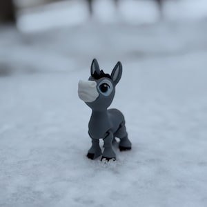 May include: A gray donkey figurine with a white face and blue eyes stands on a snowy surface. The donkey is looking up and has a playful expression.