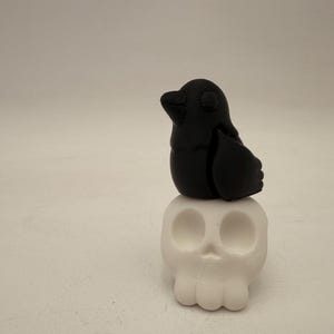 Puede incluir: Una figurita de pájaro negro posada sobre una calavera de cerámica blanca. El pájaro tiene un cuerpo redondeado y un pequeño pico. La calavera tiene dos cuencas de los ojos y una pequeña boca.