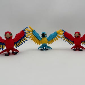 Puede incluir: Tres loros de juguete con las alas extendidas. Los loros son rojos, azules y amarillos. Los loros rojos tienen plumas amarillas y azules en las alas. El loro azul tiene plumas amarillas en las alas.