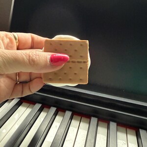 May include: A hand holding a graham cracker sandwich with white frosting between the crackers. The sandwich is being held over a black and white keyboard.