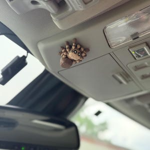 May include: A brown and tan spider-shaped car accessory, possibly an air freshener or decorative item, is affixed to the interior ceiling of a vehicle. The spider has a round body and multiple legs, with a detailed, textured appearance. The car's interior is light-colored.