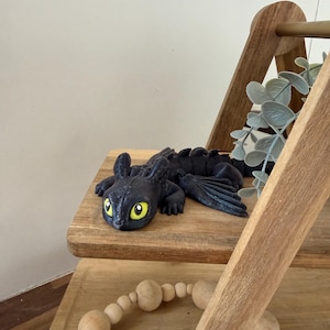 May include: A black dragon figurine with yellow eyes and detailed wings rests on a wooden shelf. The dragon is lying down, with its wings spread slightly. The shelf is part of a wooden decorative piece, with a strand of wooden beads below.