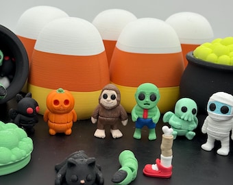 Peut inclure: Une collection de petites figurines imprimées en 3D comprenant une citrouille, une créature brune, un zombie vert, une figure ressemblant à une pieuvre, une momie blanche, un chat noir, une jambe coupée et un ver vert devant des maïs sucrés.