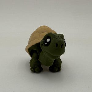 以下が含まれることがあります： 茶色の甲羅と大きな黒い目をした、小さな緑色の亀の置物。亀は微笑んでおり、3Dプリントされた質感があります。