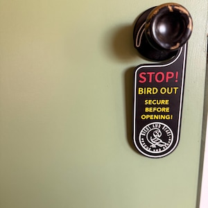 May include: A black door hanger with white text that reads "STOP! BIRD OUT SECURE BEFORE OPENING!" The hanger has a logo of a bird and the words "BIRDS AND STARS RESCUE AND STARS". The door is a light green colour.