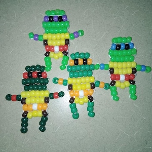 May include: Four handmade Teenage Mutant Ninja Turtle figures made from colourful beads. Each turtle has a different coloured mask: purple, blue, red, and orange. The figures are green, yellow, and brown, with arms and legs extended.