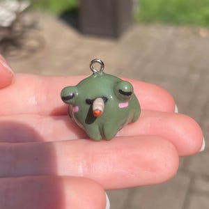 May include: A small, green frog charm with a silver-colored loop for hanging. The frog has black eyes, pink cheeks, and a small, brown object in its mouth. The charm is held in a person's hand.