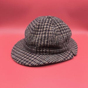 May include: A brown and black patterned hat with a rounded crown and a short brim. The hat features a classic houndstooth design, offering a vintage aesthetic. The hat is set against a solid red background.