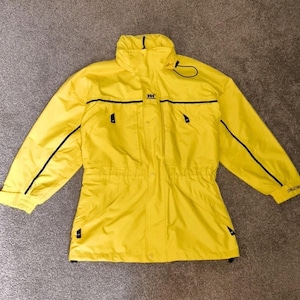 May include: Bright yellow jacket with black trim and the "HH" logo. The jacket has a high collar, a drawstring waist, and multiple pockets. The sleeves have black accents and the cuffs are adjustable. A practical and stylish outerwear piece.