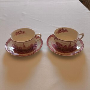 May include: Two ceramic teacups and matching saucers featuring a deep red transferware pattern. The design depicts a landscape with a church spire and trees, with floral motifs inside the rims. The cups have a rounded shape and side handles.