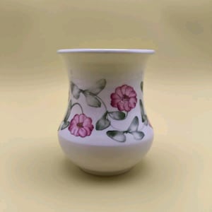 Può includere: Un vaso di ceramica bianco con base arrotondata e parte superiore leggermente svasata. Il vaso è decorato con fiori rosa e foglie verdi. Il motivo floreale è dipinto attorno al corpo del vaso.