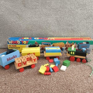 Pode incluir: Um conjunto de comboio de mercadorias de brinquedo de madeira com vários vagões e blocos coloridos. O conjunto inclui um vagão azul com a etiqueta "blocks", um vagão amarelo com "shell" impresso e uma locomotiva com a parte superior verde. O conjunto está numa superfície castanha.