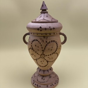 May include: A light brown, lidded urn with a pedestal base. The urn features darker brown accents and a floral design. It has two small handles and a pointed finial on the lid. The base has a decorative pattern.