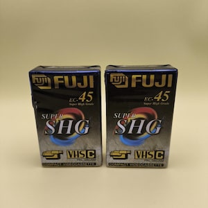 Peut inclure: Deux cassettes vidéo compactes FUJI EC-45 Super High Grade VHS-C. Les boîtes sont noires avec des accents dorés et rouges. Le texte "SUPER SHG" est bien en évidence, ainsi que le logo VHS-C.