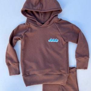 May include: A brown hooded sweatshirt and matching shorts set for a child. The sweatshirt has a blue wave graphic on the front.