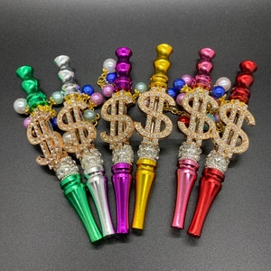 May include: Six colorful metal smoking pipes with a dollar sign design and rhinestones. The pipes are in shades of green, silver, purple, gold, pink, and red.