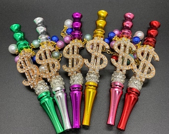 May include: Six colourful metal smoking pipes with a dollar sign design and rhinestones. The pipes are in shades of green, silver, purple, gold, pink and red.