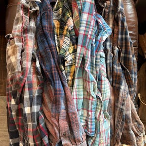 May include: A collection of plaid flannel shirts in various colors, including red, blue, green, and brown. The shirts are button-down with long sleeves, and are folded or draped together. The fabric appears to be soft and worn.