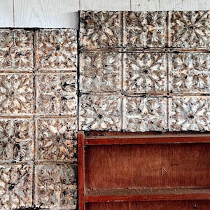 May include: Decorative, vintage-style metal ceiling tiles with a floral pattern in shades of white, brown, and gray. The tiles are arranged in a grid and rest above a dark brown wooden shelf. The background is a light-colored wooden wall.