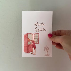 May include: A rectangular greeting card featuring a watercolour illustration of a person in a red polka-dot outfit leaning into an open red refrigerator. The text 'Heiße Grüße' is written in brown script above the refrigerator, and a small white fan stands to the right.