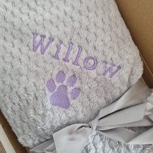 May include: A soft, gray, plush blanket with a purple paw print and the name "Willow" embroidered on it. The blanket is tied with a gray ribbon.