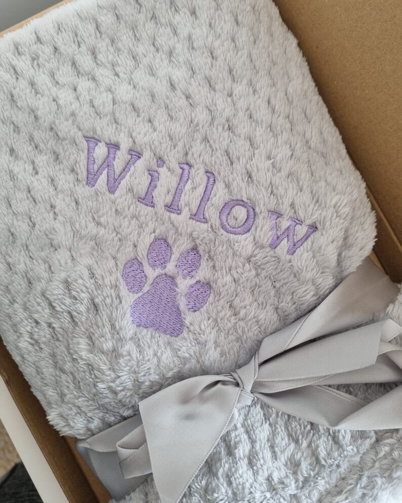 May include: A soft, gray, plush blanket with a purple paw print and the name "Willow" embroidered on it. The blanket is tied with a gray ribbon.