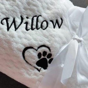 Puede incluir: Una manta blanca y afelpada para mascotas bordada con el nombre "Willow" en escritura negra. Un diseño de huella de pata en forma de corazón negro está debajo del nombre. Una cinta de satén blanco está atada en un lazo en el lado derecho.