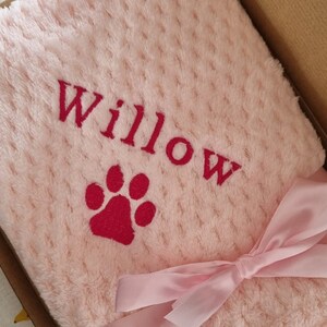 May include: A pink plush blanket with a red paw print and the name "Willow" embroidered on it. The blanket is tied with a pink ribbon.