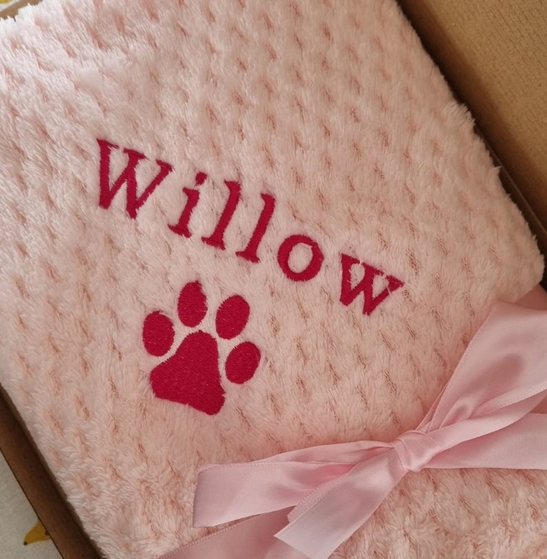 May include: A pink plush blanket with a red paw print and the name "Willow" embroidered on it. The blanket is tied with a pink ribbon.