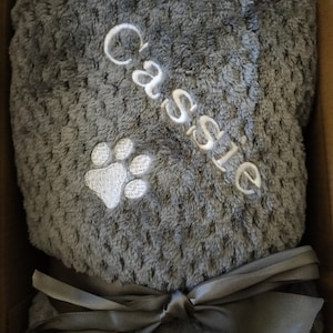 May include: A gray plush blanket with a white paw print and the name "Cassie" embroidered on it. The blanket is tied with a gray satin ribbon.