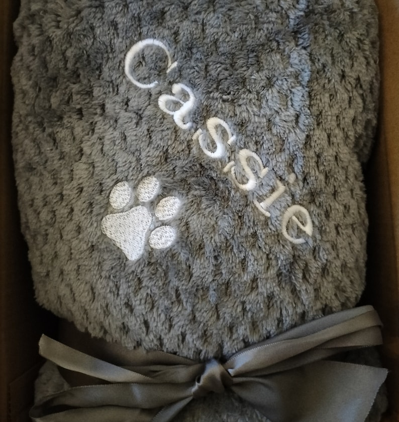 May include: A gray plush blanket with a white paw print and the name "Cassie" embroidered on it. The blanket is tied with a gray satin ribbon.