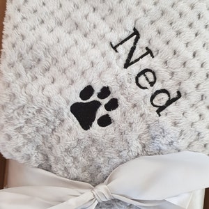 May include: A gray and white plush blanket with a black paw print and the name "Ned" embroidered on it. The blanket is tied with a white ribbon.