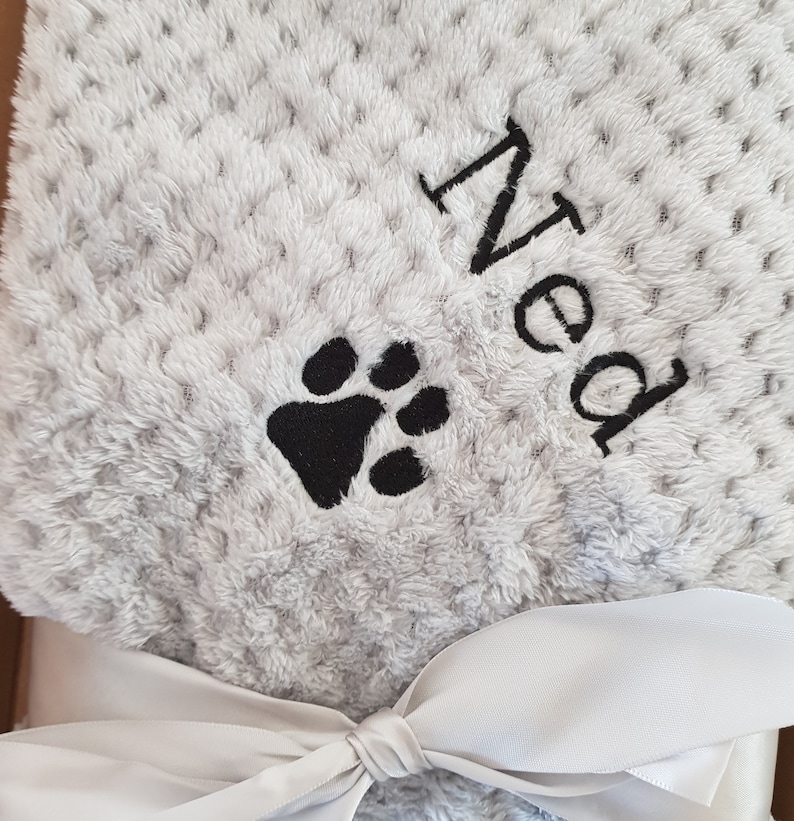 May include: A gray and white plush blanket with a black paw print and the name "Ned" embroidered on it. The blanket is tied with a white ribbon.